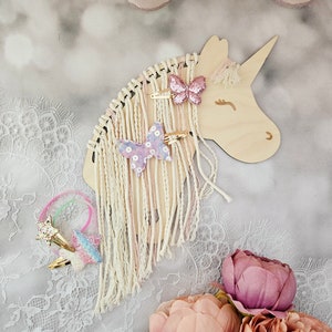 K&ouml;nnte beinhalten: Holzhalter f&uuml;r Haarschmuck in Form eines Einhorns mit wei&szlig;em Fransenbesatz und rosa und violetten Schmetterlingshaarklammern. Der Halter befindet sich auf einer wei&szlig;en Spitze mit rosa Blumen.