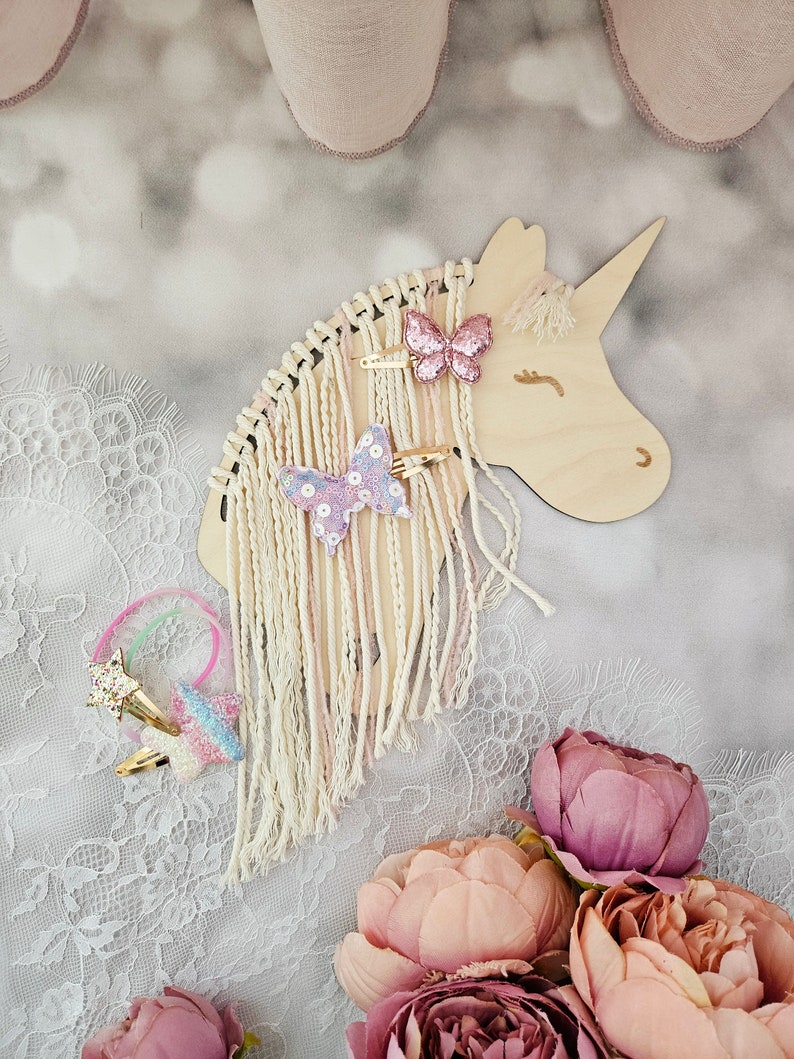 K&ouml;nnte beinhalten: Holzhalter f&uuml;r Haarschmuck in Form eines Einhorns mit wei&szlig;em Fransenbesatz und rosa und violetten Schmetterlingshaarklammern. Der Halter befindet sich auf einer wei&szlig;en Spitze mit rosa Blumen.