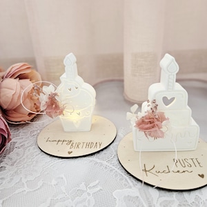 May include: Two white ceramic cake-shaped candle holders with heart cutouts and a single candle. Each sits on a wooden base with text that reads "happy BIRTHDAY" and "Kuchen PUSTE". Pink flowers and fairy lights are attached.