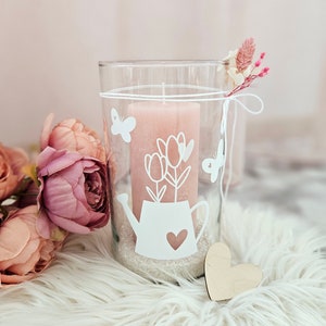 Pu&ograve; includere: Un portacandele in vetro trasparente con una candela rosa all'interno. Il portacandele ha un design bianco a forma di annaffiatoio con fiori e farfalle. Il portacandele &egrave; appoggiato su una superficie bianca e soffice.