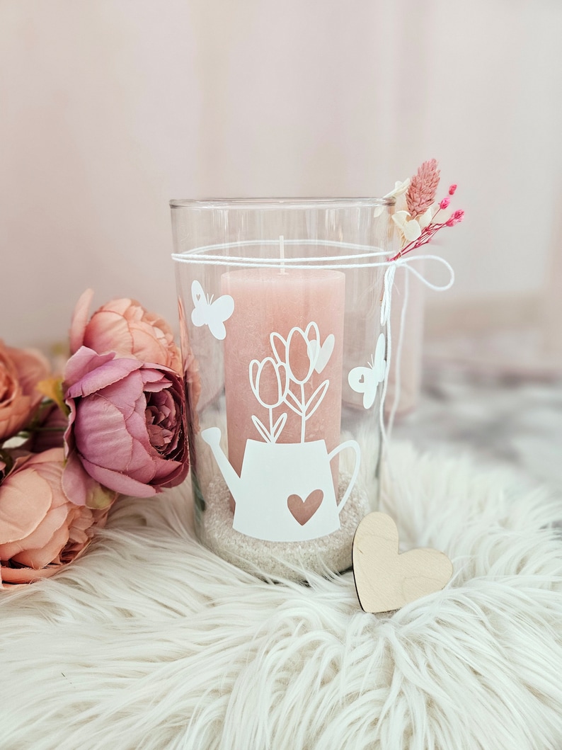 Pu&ograve; includere: Un portacandele in vetro trasparente con una candela rosa all'interno. Il portacandele ha un design bianco a forma di annaffiatoio con fiori e farfalle. Il portacandele &egrave; appoggiato su una superficie bianca e soffice.