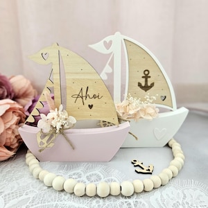 Op de afbeelding: Twee decoratieve houten zeilboten, één roze en één wit, met bloemaccenten. De roze boot heeft het woord "Ahoi" op het zeil. Een houten kralenkrans en een klein ankeraccent maken het nautische thema compleet.