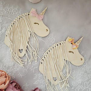 K&ouml;nnte beinhalten: Zwei Wandbeh&auml;nge aus Holz in Form eines Einhornkopfes mit Garnfransen. Das Einhorn links hat eine rosa Schleife und den Namen "Selena" darauf geschrieben. Das Einhorn rechts hat eine rosa und gelbe Schleife und den Namen "Nellie" darauf geschrieben.