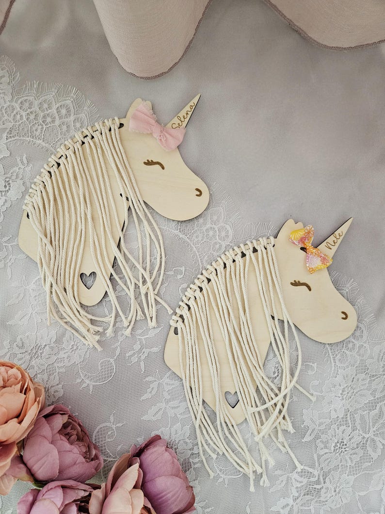 K&ouml;nnte beinhalten: Zwei Wandbeh&auml;nge aus Holz in Form eines Einhornkopfes mit Garnfransen. Das Einhorn links hat eine rosa Schleife und den Namen "Selena" darauf geschrieben. Das Einhorn rechts hat eine rosa und gelbe Schleife und den Namen "Nellie" darauf geschrieben.