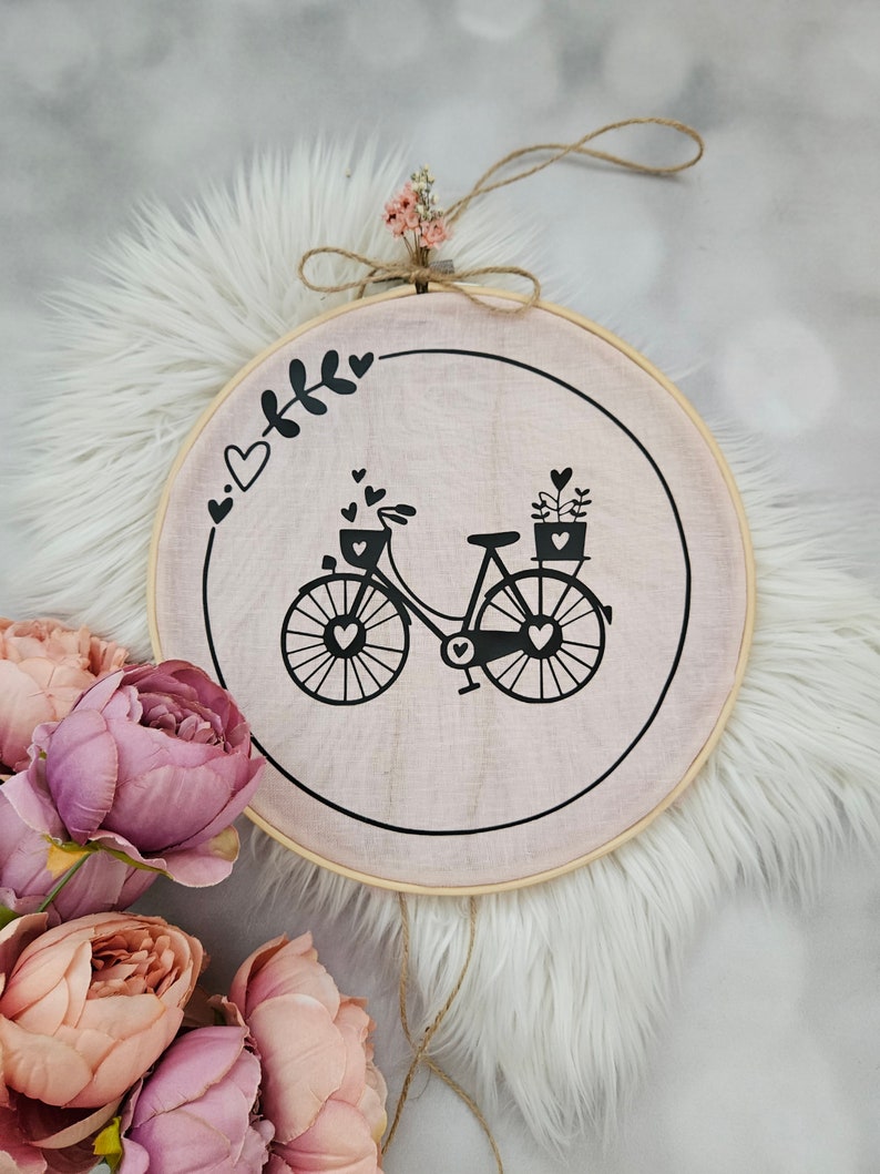 Pode incluir: Um bastidor de bordado redondo de madeira com tecido rosa apresentando um desenho de bicicleta preta com cora&ccedil;&otilde;es. O bastidor &eacute; adornado com um pequeno buqu&ecirc; de flores secas e um la&ccedil;o de barbante para pendurar. Flores rosa e p&ecirc;ssego em primeiro plano. O di&acirc;metro &eacute; de cerca de 20 cm.