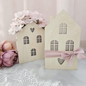 Könnte beinhalten: Zwei Holzhäuser mit ausgeschnittenen Fenstern und Herzen. Ein Haus ist mit rosa Blumen und das andere mit einem rosa Band verziert.