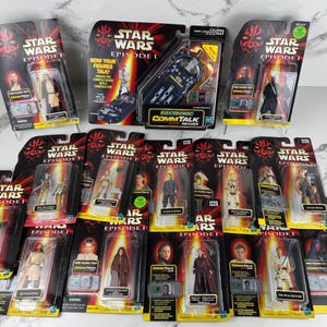 Könnte beinhalten: Eine Sammlung von Star Wars Episode I Actionfiguren in ihrer Originalverpackung. Die Figuren werden auf einer weißen Oberfläche präsentiert. Die Verpackung zeigt Charakterbilder und das Star Wars-Logo.