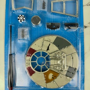 Puede incluir: Kit de modelo de nave espacial, presentado en una bandeja de plástico azul. El kit incluye un cuerpo principal circular con paneles de colores, una ventana de cabina central y varios componentes más pequeños, además de un destornillador.