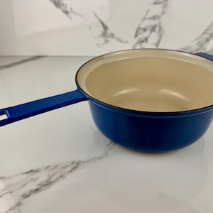 Puede incluir: Una cacerola azul de hierro fundido esmaltado con un mango largo. El interior es de color crema y el borde es negro. El mango tiene un agujero para colgar. Esta olla es adecuada para su uso en la estufa.