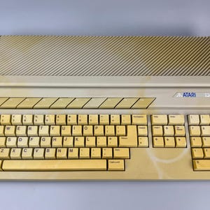 Könnte beinhalten: Ein beigefarbener Vintage-Computer Atari 1040ST mit einer vollständigen QWERTY-Tastatur inklusive Ziffernblock, einer Reihe von Funktionstasten und einem oberen Lüftungsgitter. Das Kunststoffgehäuse weist Vergilbungen und Verfärbungen auf.