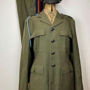 May include: Olive green military uniform consisting of a jacket, trousers, and a peaked cap. The jacket has four front pockets and black buttons. The cap is white with a red band and a gold emblem. The trousers have a colorful stripe.