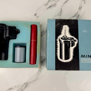 Może przedstawiać: Zestaw do wywoływania w świetle dziennym Minox w stylu vintage. Zestaw zawiera czarny zbiornik wywoływaczy, szarą kasetę na film i czerwony pojemnik. Zestaw jest w oryginalnym pudełku z logo i tekstem Minox.