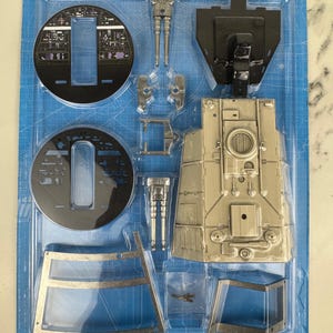 May include: A model kit of a spacecraft, featuring various unassembled parts in a blue plastic tray. The kit includes components in silver and black, such as cockpit sections, engine parts, and structural elements. The tray has a blueprint-style design.