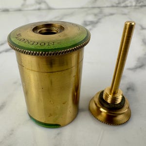 Op de afbeelding: Een vintage messing inktpot met bijpassende penhouder. De inktpot heeft een groene rand met het woord "LONDON" erop gedrukt. De penhouder is een lange, dunne staaf met een cirkelvormige basis. Ongeveer 6 cm hoog.