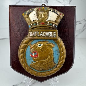 May include: A wooden plaque with a shield shape, featuring a detailed emblem. The emblem includes a crown, the word "IMPLACABLE", and a roaring lion. The plaque is dark brown with a glossy finish, and the emblem is painted in gold, blue, red, and white.