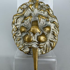 May include: A circular brass door knocker featuring a detailed lion head relief with a prominent mane and a long, pointed tongue extending downwards. The metal surface displays a mix of polished gold-toned highlights and darker, recessed areas.