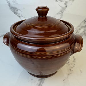 Op de afbeelding: Een bruine keramische braadpan met een deksel. De schaal heeft twee handvatten en een decoratief stippenpatroon rond de rand en het deksel. Het deksel heeft een kleine knop bovenop.