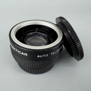 Op de afbeelding: Een zwarte Prakticar Auto Teleconverter camera-lensaccessoire met een zilveren metalen montagering en een losse zwarte ronde lensdop. De zijkant van de zwarte behuizing is voorzien van witte tekst met de tekst Prakticar en Auto Teleconverter.