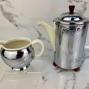 May include: A vintage chrome coffee pot and creamer set. The coffee pot has a hammered metal finish, a cream-colored handle and spout, and a dark brown knob on the lid. The creamer has a matching hammered metal finish and cream-colored handle and spout.