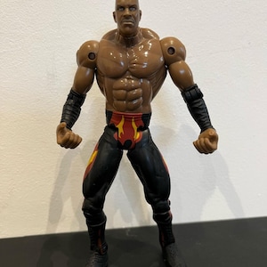 May include: A brown action figure of a muscular man in a wrestling pose. The figure wears black and red wrestling attire, including boots and wristbands. The figure has a stern expression and is posed with arms slightly bent.