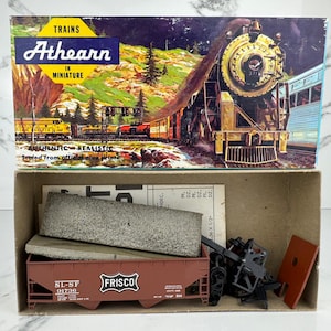 May include: Athearn model train set box with a detailed illustration of a steam engine and train cars. The box is open, revealing a brown Frisco train car, a gray stone-like piece, and other model train parts. The box reads "TRAINS in MINIATURE".