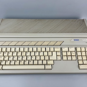 Könnte beinhalten: Ein beigefarbener Atari 520ST Computer mit einer vollständigen QWERTY-Tastatur inklusive Ziffernblock, einer Reihe von Funktionstasten und einem großen, geneigten Lüftungsgitter an der Oberseite. Das Atari-Logo und der Modellname sind rechts blau aufgedruckt.