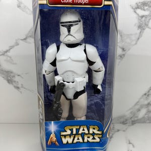 Pode incluir: Uma figura de ação Star Wars Clone Trooper em uma caixa de exibição de plástico transparente. A figura é branca com detalhes em preto e segura um blaster preto. A caixa tem o texto "ATTACK OF THE CLONES" e "Clone Trooper". O logotipo Star Wars também é visível.