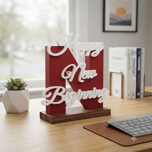 Op de afbeelding: Een donkerrode houten letter N op een rechthoekige bruine voet, voorzien van witte cursieve letters met de tekst Happy New Beginning. Het decoratieve object staat op een bureau naast een kleine vetplant en enkele boeken.