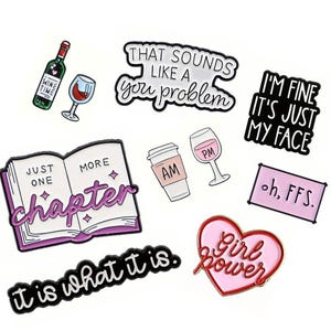 May include: A selection of enamel pins with diverse designs and text. The pins feature a wine bottle and glass, a coffee cup, a book with the text "Just One More Chapter", a heart with "Girl Power", and phrases like "It is what it is."