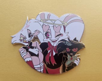 May include: A white, pink, and red enamel pin featuring three cartoon characters. The characters are a white rabbit with pink ears and a halo, a black cat with green eyes, and a white rabbit with pink ears and a red bow.