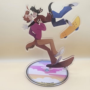 Könnte beinhalten: Ein durchsichtiger Acryl-Ständer mit einem bunten Sonnenuntergangs-Hintergrund. Auf dem Ständer sind zwei Comicfiguren abgebildet, eine ist ein brauner Hund mit einem roten Shirt und die andere ist ein schwarzer Hund mit einem blauen Shirt. Beide Figuren halten Skateboards.