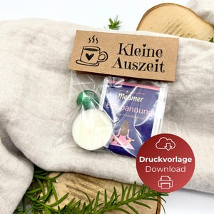 May include: A small gift set with a candle, tea bag, and a card that reads "Kleine Auszeit". The candle is white with a green top. The tea bag is blue and white. The gift set is on a beige cloth.