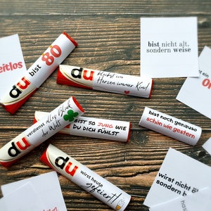 Pu&ograve; includere: Una collezione di caramelle avvolte in bianco e rosso con testo in tedesco, sparse su una superficie di legno. Le caramelle presentano frasi come "du bist 80" e "bist so jung wie du dich f&uuml;hlst". Sono presenti anche piccole carte bianche con testo.