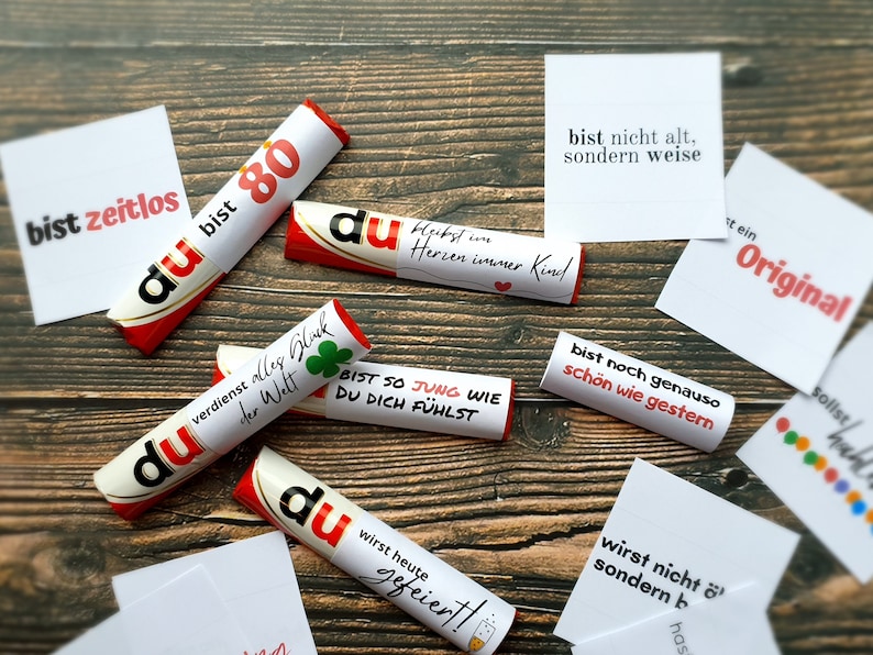 Pu&ograve; includere: Una collezione di caramelle avvolte in bianco e rosso con testo in tedesco, sparse su una superficie di legno. Le caramelle presentano frasi come "du bist 80" e "bist so jung wie du dich f&uuml;hlst". Sono presenti anche piccole carte bianche con testo.