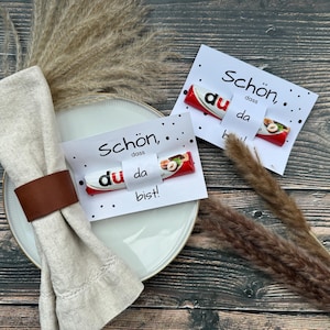 May include: Two white cards with black text and black dots. The text on the cards reads "Schon, dass du da bist!"  Each card has a chocolate bar with a red wrapper and a hazelnut on top.
