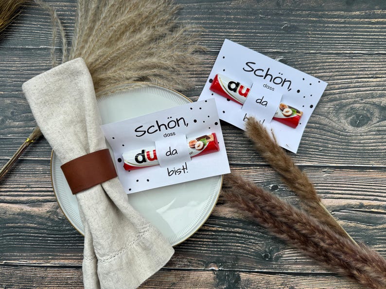 May include: Two white cards with black text and black dots. The text on the cards reads "Schon, dass du da bist!"  Each card has a chocolate bar with a red wrapper and a hazelnut on top.