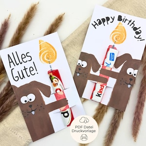 May include: Two greeting cards with cartoon dog illustrations. One card says "Alles Gute!" and the other says "Happy Birthday!". Each card features a candy bar designed to look like a candle with a flame.