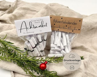 May include: Two small cellophane bags filled with rolled white paper, each with a decorative label. One label reads "Achtsamkeit" and the other "Adventskalender". A sprig of green foliage and a red ornament are in the foreground.