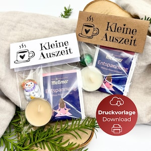 以下が含まれることがあります： 「Kleine Auszeit」の文字とコーヒーカップのイラストが入った小さなギフトバッグが2つ。各バッグにはお茶、キャンドル、小さな装飾品が入っています。バッグは、緑とベージュの布の背景に、木の輪切りに置かれています。