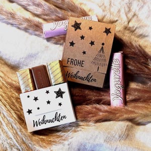 May include: A festive arrangement featuring small chocolate bars in a white box decorated with black stars and the word "Weihnachten". A brown box with "Frohe Weihnachten" and a Christmas tree is also present, along with pink and purple candy bars.
