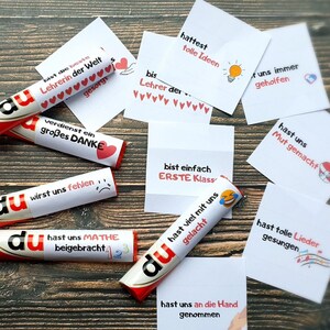 May include: A collection of white paper labels and candy bars wrapped in white paper, each featuring German text and small colorful icons. The labels include phrases like bist die beste Lehrerin der Welt, hattest tolle Ideen, and hast uns Mathe beigebracht.