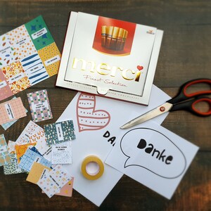 May include: A box of Merci Finest Selection assorted chocolates with a red heart and the word "merci" in gold lettering. The box is on a wooden surface with a variety of colorful paper scraps and a white card with the word "Danke" in black lettering.