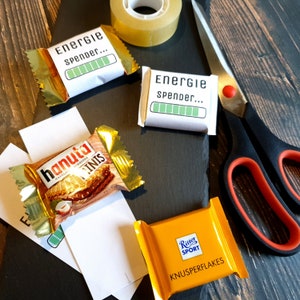 May include: A flat lay of various items on a wooden surface. There are two white boxes with the text "Energie spender..." and a green progress bar. There is a chocolate bar with the text "hanuta Minis" and a yellow chocolate bar with the text "Ritter Sport Knusperflakes". There is also a roll of tape and a pair of red-handled scissors.