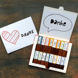 May include: A box of assorted chocolate bars with colorful wrappers and the word "Danke" printed on the front. The box is open and the chocolate bars are visible. There is a card with a heart and the word "Danke" on it.