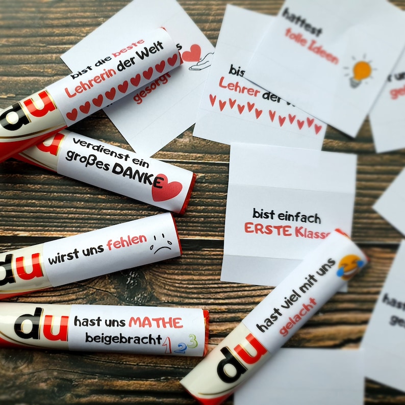 May include: Several cylindrical chocolate bars wrapped in white paper labels with German text and red heart icons, accompanied by small white square cards featuring printed messages, lightbulb graphics, and emojis, arranged in a scattered layout.