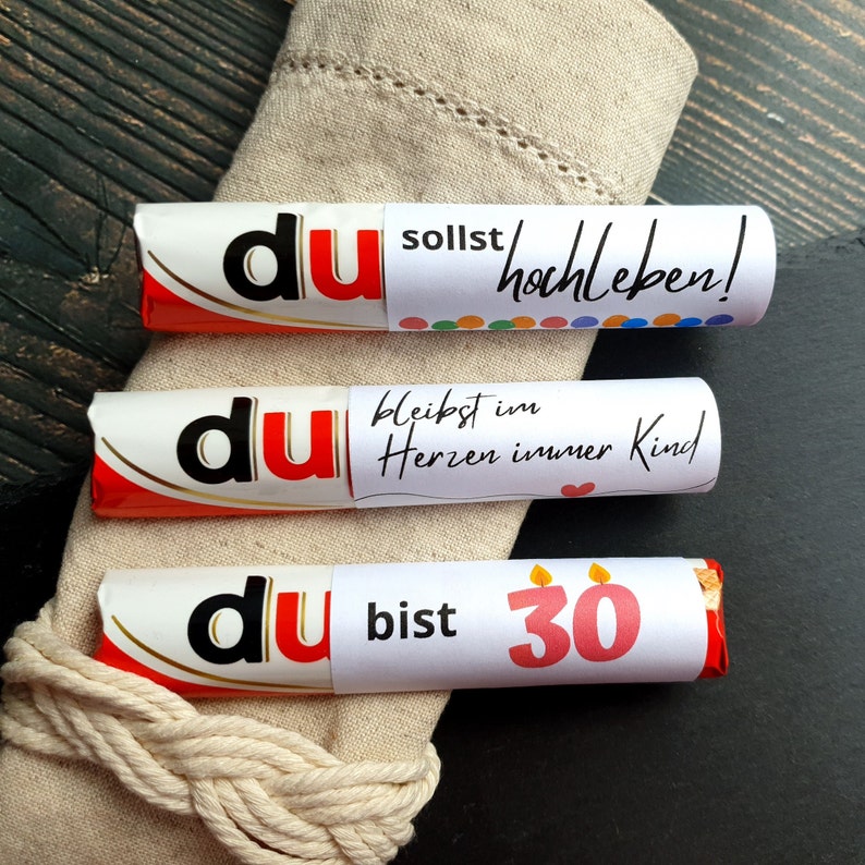 May include: Three chocolate bars wrapped in red and white foil with personalized messages. The top bar says "du sollst hochleben!" The middle bar says "du bleibst im Herzen immer Kind." The bottom bar says "du bist 30".