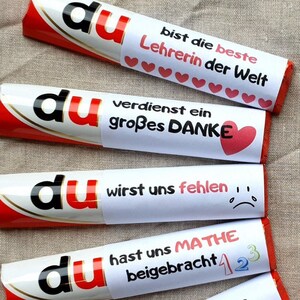 May include: Four chocolate bars wrapped in white paper with red accents. Each bar features the word du in black and red letters, followed by German text: bist die beste Lehrerin der Welt with hearts, verdienst ein gro&szlig;es DANKE with a heart, wirst uns fehlen with a sad face, and hast uns MATHE beigebracht with numbers 1, 2, and 3.