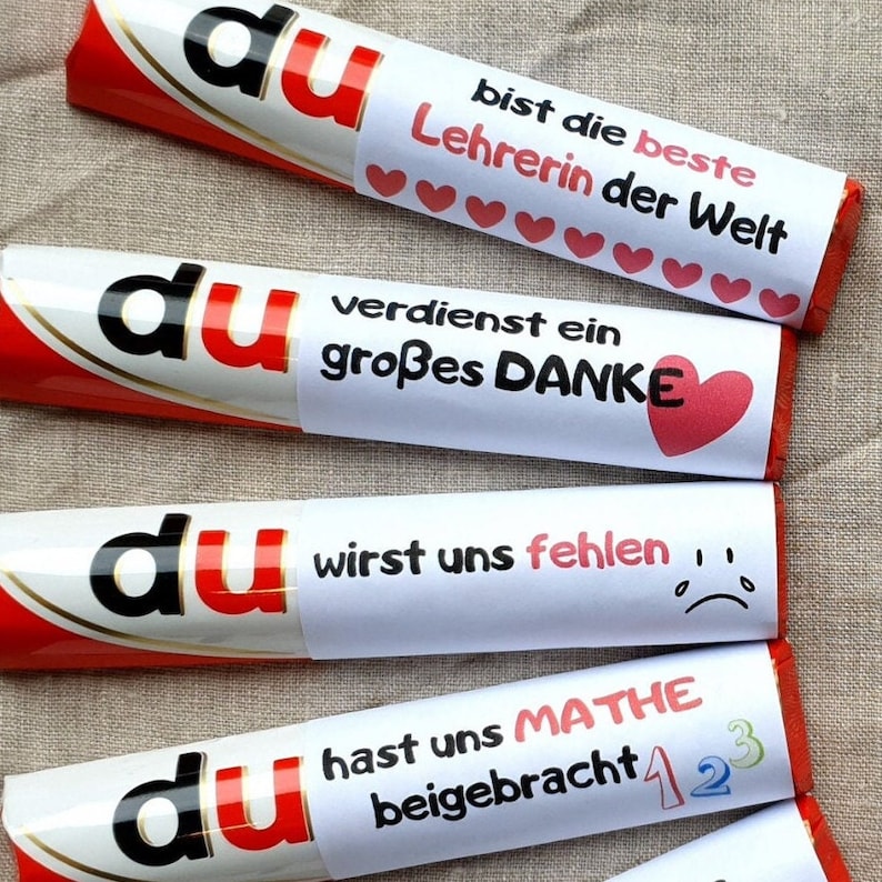 May include: Four chocolate bars wrapped in white paper with red accents. Each bar features the word du in black and red letters, followed by German text: bist die beste Lehrerin der Welt with hearts, verdienst ein gro&szlig;es DANKE with a heart, wirst uns fehlen with a sad face, and hast uns MATHE beigebracht with numbers 1, 2, and 3.