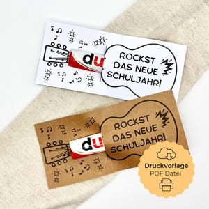 Könnte beinhalten: Zwei Karten mit einer Gitarrenillustration und dem Text "ROCKST DAS NEUE SCHULJAHR!". Eine Karte ist weiß, die andere braun. Ein rot-weißer Schokoriegel ist in jede Karte eingesetzt. Noten und Sterne verzieren die Karten.