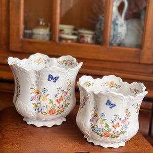 May include: Two white ceramic planters with scalloped rims and embossed detailing. Each planter features a colourful floral pattern with orange, blue, and yellow flowers, green foliage, and a prominent blue butterfly on the front surface.